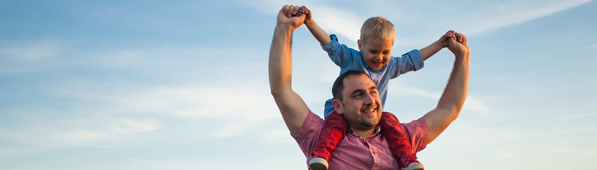 Man with his son on his shoulders holding his son's hands up in the air in an open field with blue skies.