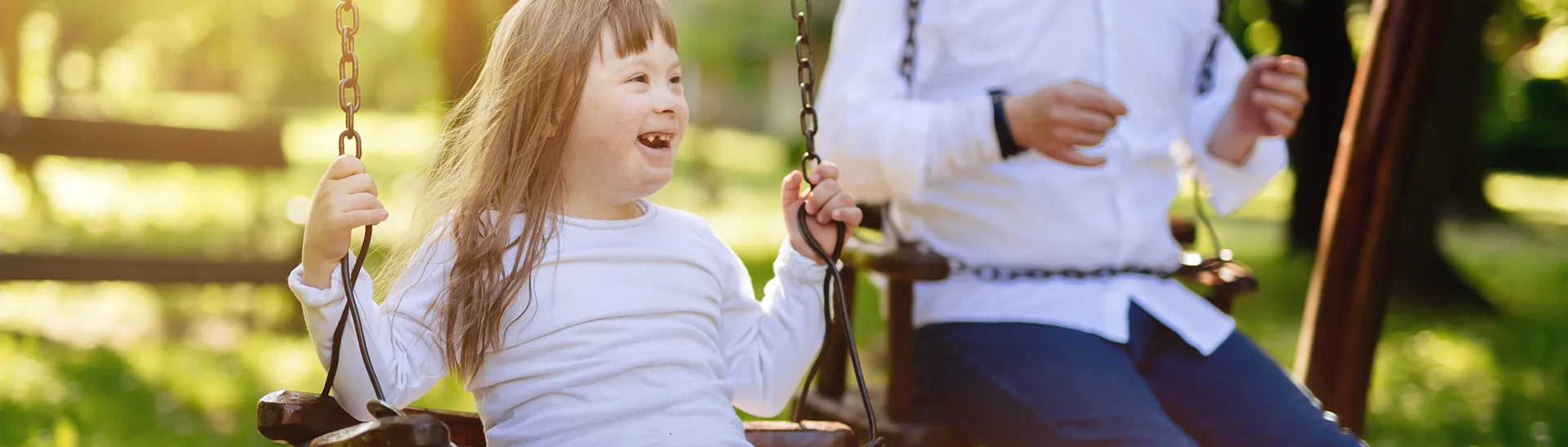 A little girl with down syndrome is swinging on a playground with another older boy swinging next to her.