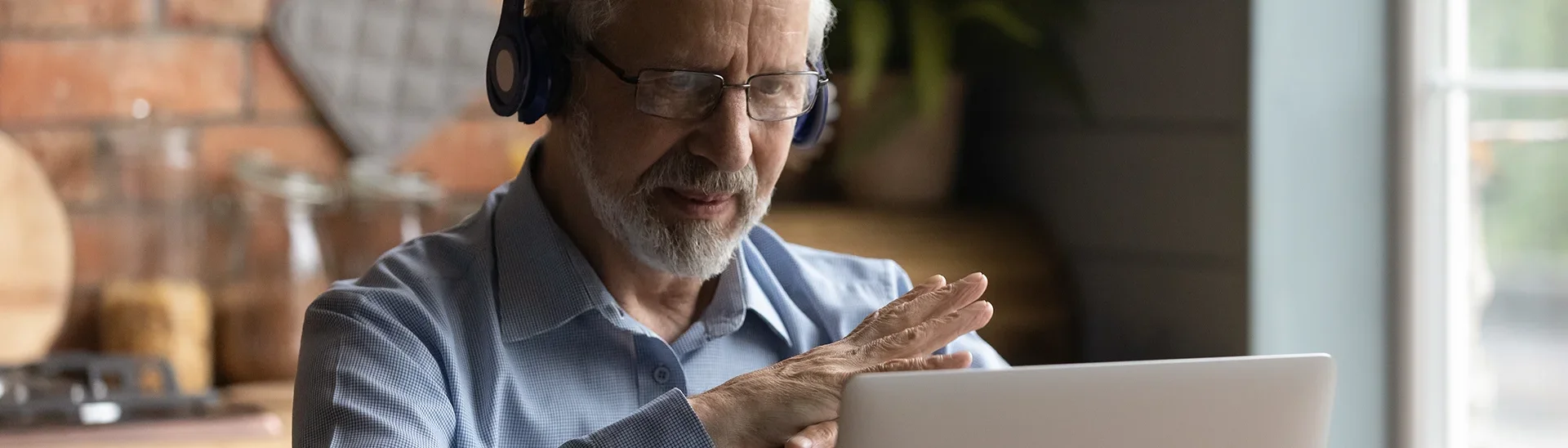 Hombre mayor con gafas y auriculares puestos proporcionando ayuda de tutoría virtualmente con las manos cruzadas delante de su ordenador portátil.