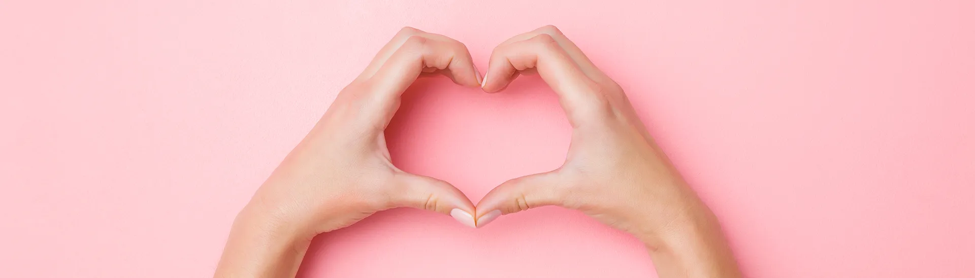 A person making the "I love you" symbol with their hands in the shape of a heart in front of a pink background.