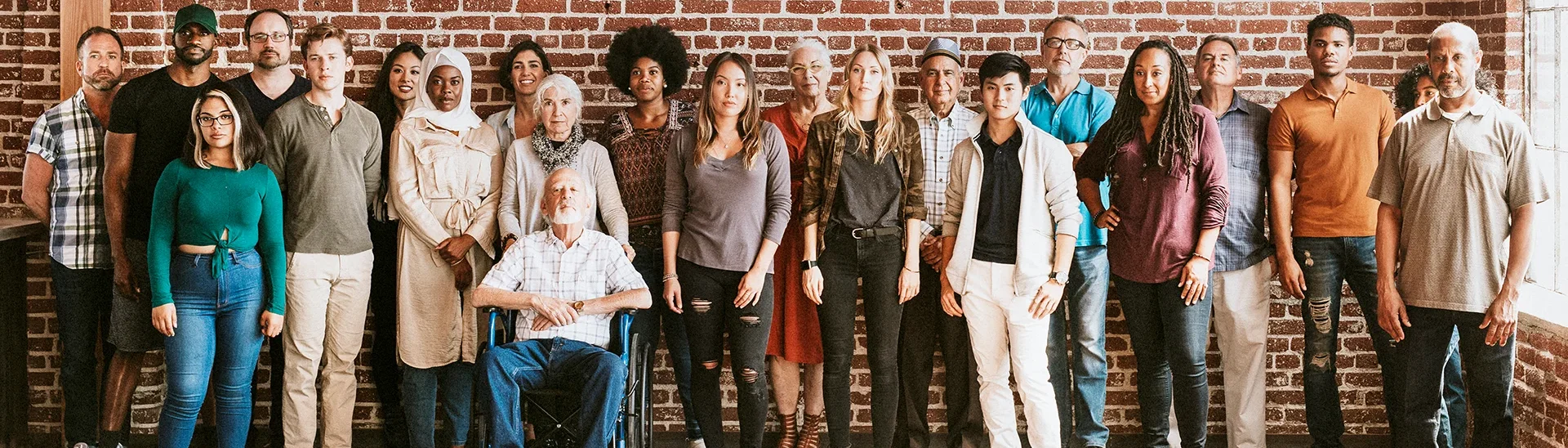 Large group of various peoples of different ethnicities and ages sitting or standing together against a brick wall.