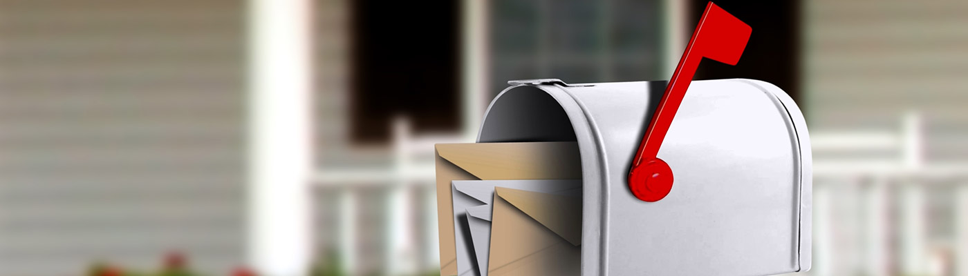 Open white mailbox with letters sticking out the front, in front of a white house with black shutters.