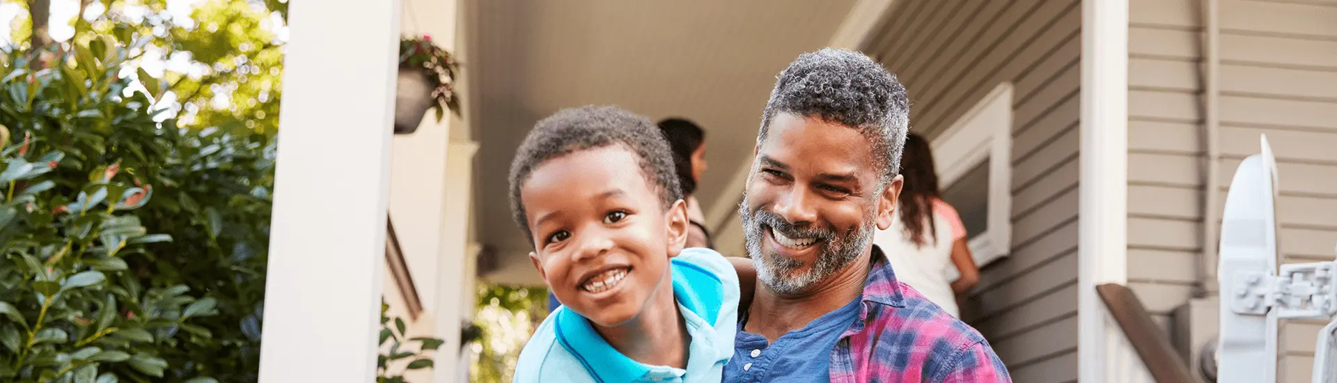 A smiling man with salt-and-pepper hair and a beard is holding a young boy in his arms. The boy, wearing a bright blue shirt, is grinning joyfully and looking at the camera. They are standing on the porch of a house with a gray exterior, white columns, and greenery in the background, giving the scene a warm and happy family atmosphere.