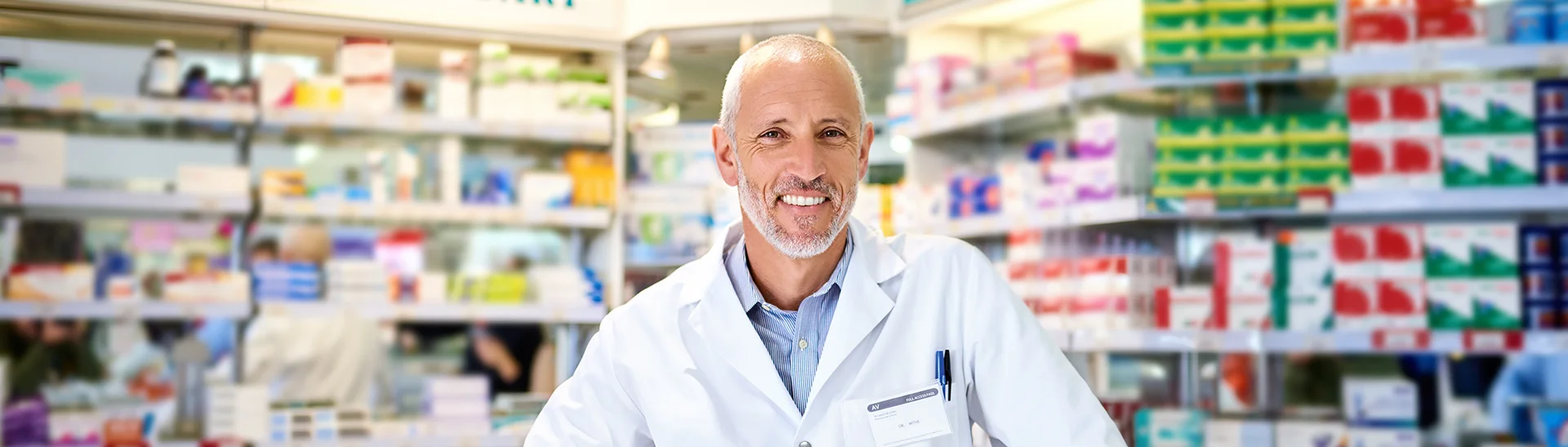 A smiling pharmacist with short gray hair and a beard is standing behind the counter of a well-stocked pharmacy. He is wearing a white lab coat and looking directly at the camera. Behind him, shelves are filled with colorful boxes of medications, health products, and signage for "Medical Aid at Main Dispensary" and "Convenient Over the Counter Medical." The pharmacy is brightly lit and organized.