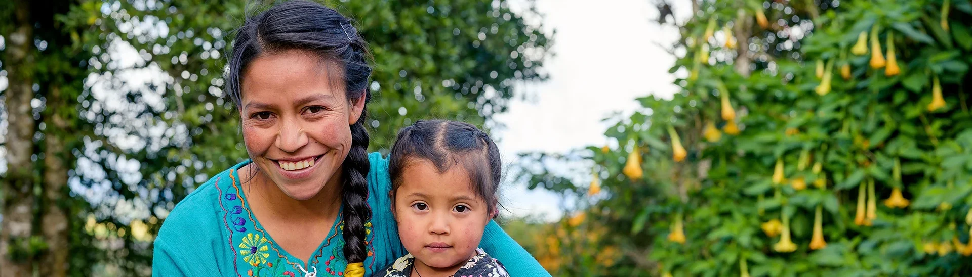 Una mujer sonriente con una blusa verde azulado con bordados de colores sostiene a una niña en brazos. La niña, vestida con un conjunto negro y rosa estampado, mira directamente a la cámara con expresión neutra. Están al aire libre, con un exuberante follaje verde y flores amarillas en forma de trompeta de fondo.