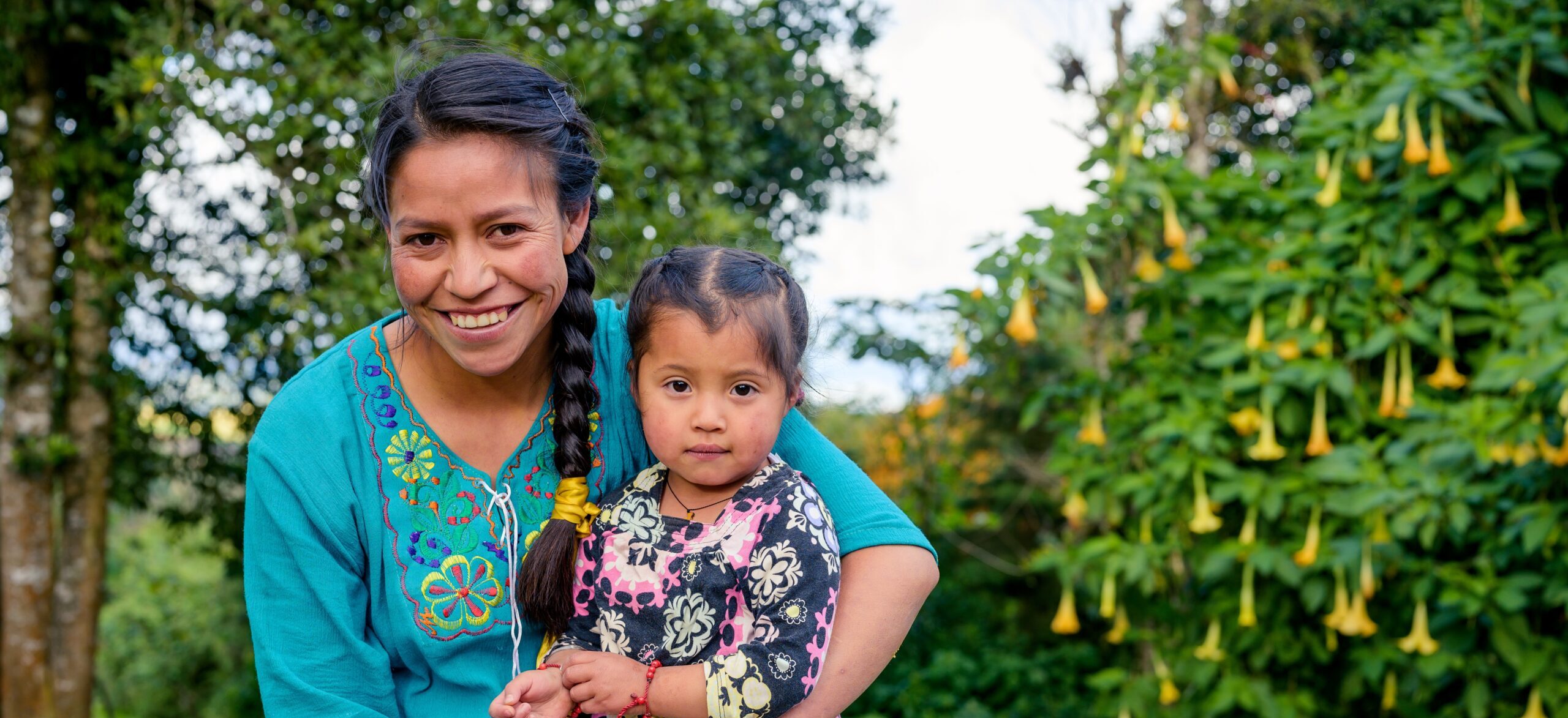 A smiling woman wearing a teal blouse with colorful embroidery holds a young girl in her arms. The girl, dressed in a black and pink patterned outfit, looks directly at the camera with a neutral expression. They are outdoors with lush green foliage and yellow trumpet-shaped flowers in the background.