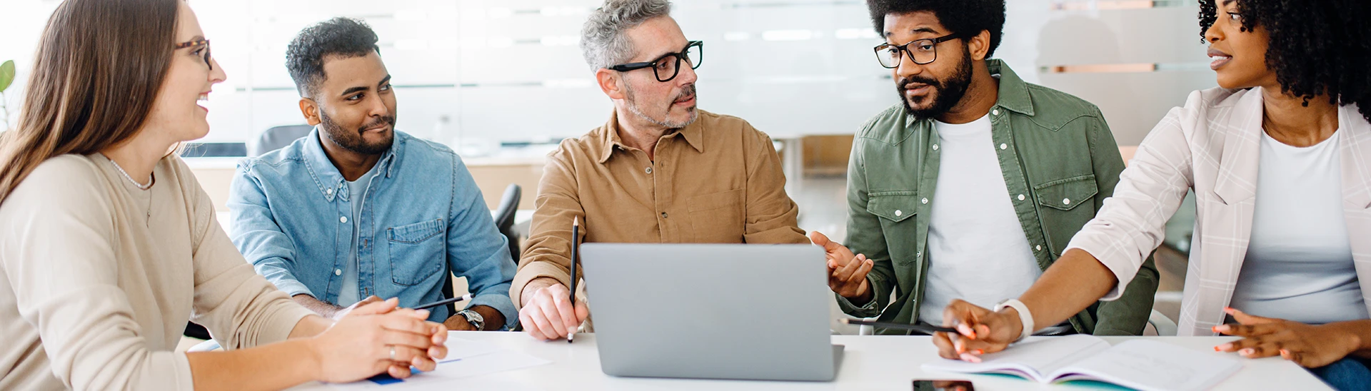 A diverse group of people are working together while sitting around a table with a single laptop open.