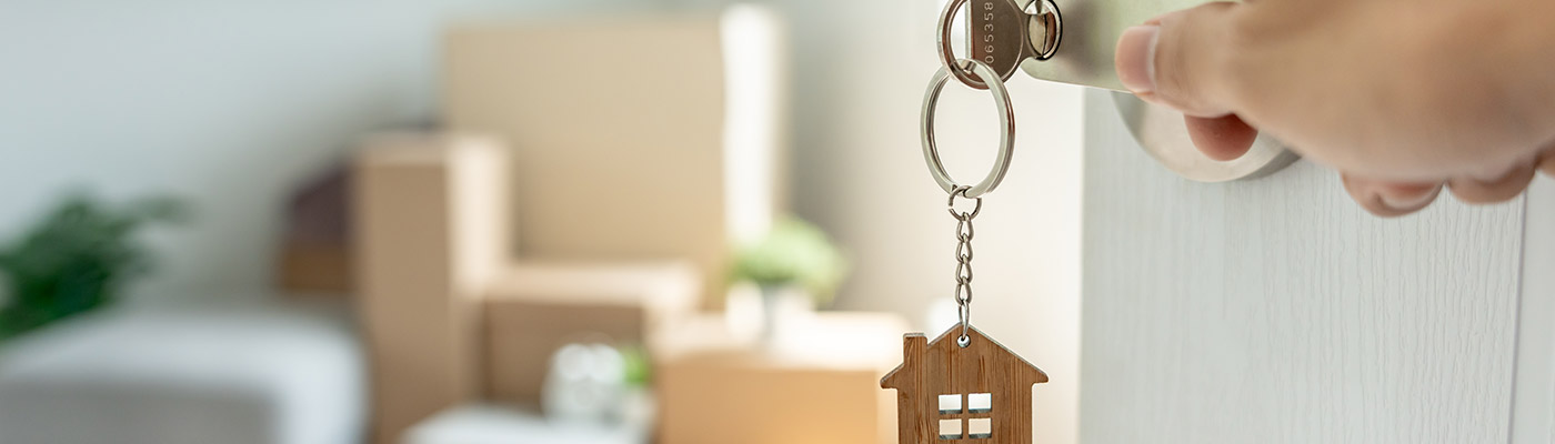 Key handing from the lock of a home's door opening to a blurry image of boxes sitting in a room.